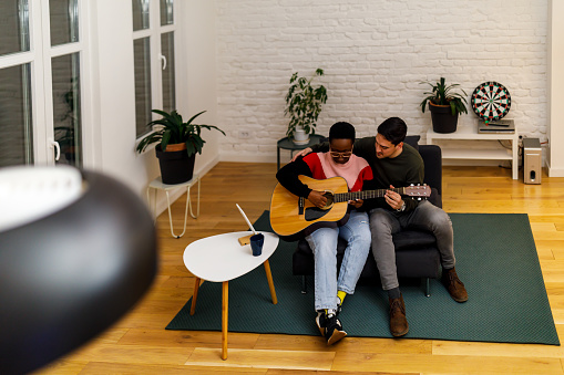 A cheerful couple is having fun at home. A young man is playing an acoustic guitar for his African wife who is enjoying the music.