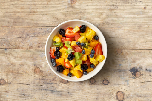 Bowl of healthy fresh summer mixed fruit salad on wooden table. Delicious and wholesome food background top view.