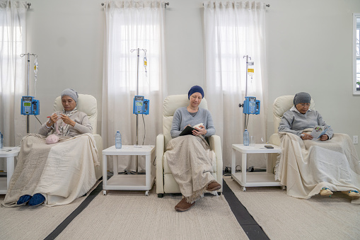 Three women sit in individual chairs as they receive their Chemotherapy by intravenous.  They are each dressed comfortably and have head scarves on to keep them warm as they engage in the activity they each brought to keep them occupied.