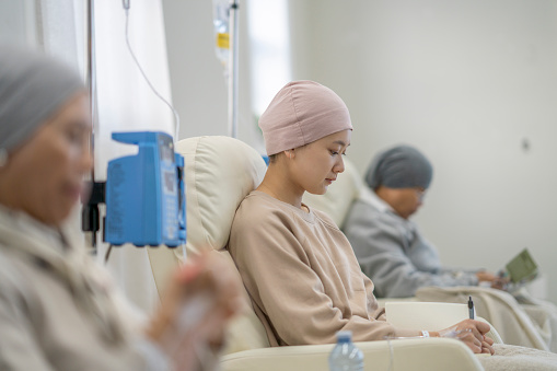 Three women sit in individual chairs as they receive their Chemotherapy by intravenous.  They are each dressed comfortably and have head scarves on to keep them warm as they engage in the activity they each brought to keep them occupied.
