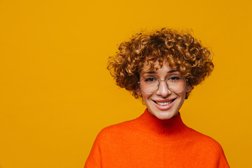 Joyful middle-aged ginger woman in eyeglasses looking at camera isolated over yellow studio background