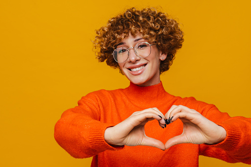 Joyful mature ginger woman in glasses make heart love gesture isolated over yellow background