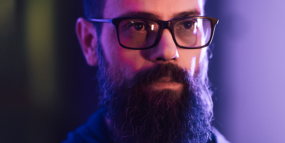 Male portrait of man with neon lighting. Red, blue, purple and magenta colours with dark background. Person wear eyeglasses, dark jacket indoors. Futuristic lifestyle concept photo with multicolour light