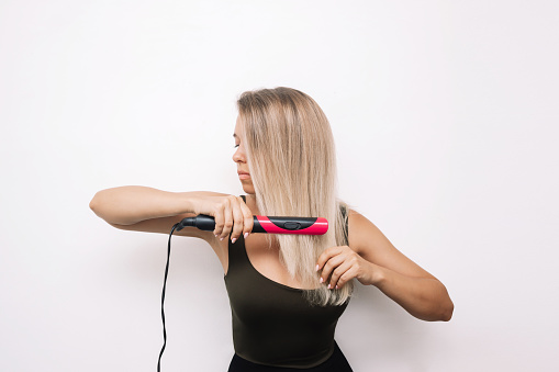 A young attractive caucasian blonde woman straightens her hair with black and pink hair straightener on a white background. Hair styling, damage