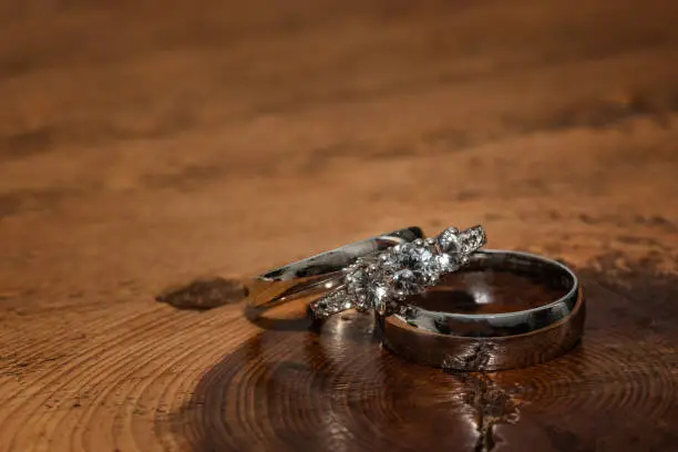 The bride engagement and wedding ring lie on top of the groom's wedding band set on a wood textured background.