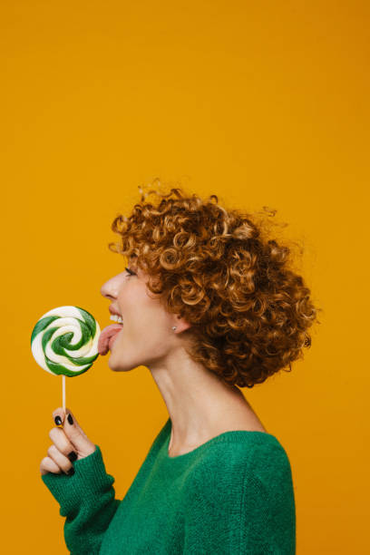 Middle-aged smiling woman liking lollipop isolated over yellow background stock photo