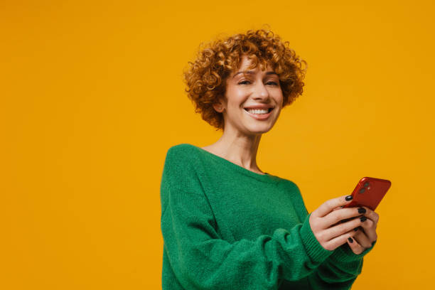 Joyful middle-aged ginger woman using smartphone isolated over yellow background stock photo