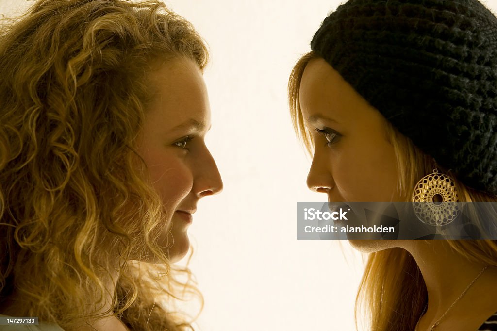 Two girls in close up looking at each other Two girls looking into each other’s eyes intently. They are very close and in profile and are not smiling. The girl on the right is wearing a black hat. The girl on the left has curly brown hair with blond streaks.  The background is bright and plain Adult Stock Photo