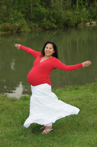 Late term pregnant woman spins around wearing white flowing skirt and red top.  Taken in countryside by a lake.