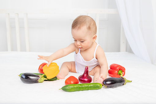 little baby sitting on the bed in the nursery with vegetables, baby food concept