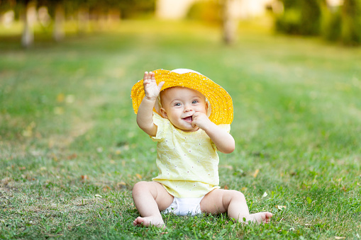 a little girl of 8 months is sitting in the summer on the green grass in a yellow summer dress and hat, a place for text.