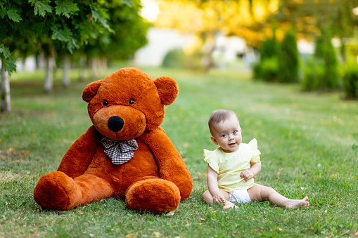 Cute little girl sitting on green grass with big Teddy bear in yellow summer dress in summer, a place for text.