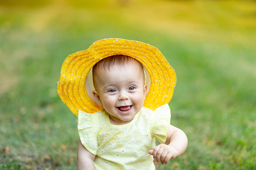 portrait of a little girl 8 months old sitting in the summer on the green grass in a yellow summer dress and hat and laughing.