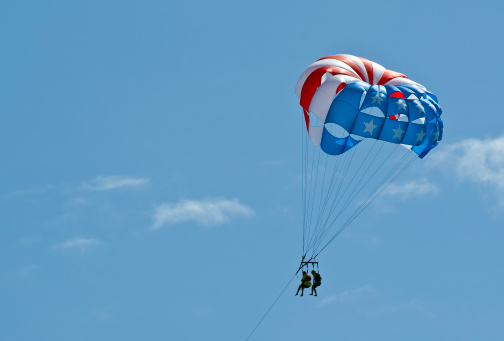 A couple of friends parasailing across a blue sky.