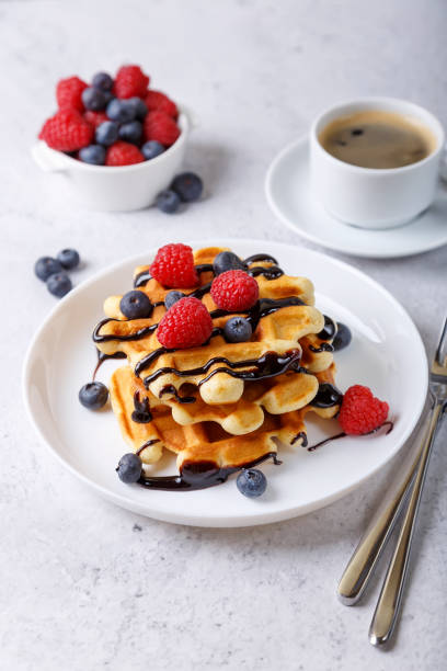 gofres vieneses o belgas con bayas frescas (frambuesas y arándanos) y salsa de chocolate en un plato blanco y una taza de café. postre tradicional. - waffle belgian waffle breakfast fruit fotografías e imágenes de stock