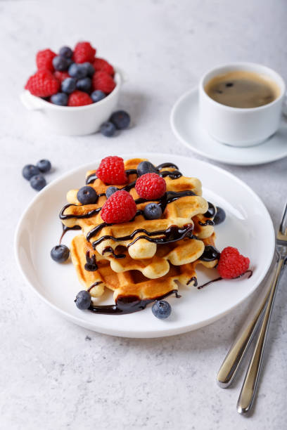 viennese or belgian waffles with fresh berries (raspberries and blueberries) and chocolate sauce on a white plate and a cup of coffee. traditional dessert. - waffle breakfast food sweet food imagens e fotografias de stock