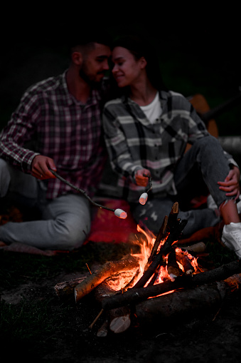 Young couple in love roasting marshmallows on campfire on nature. Lovely man and woman rest at the fire and enjoy life outdoors