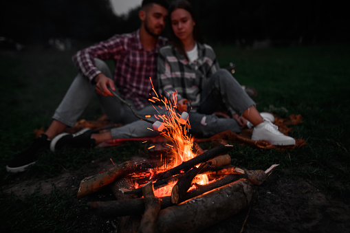 Young couple relaxing near the fire in the nature. Handsome man and beautiful woman are roasting at fire marshmallows on sticks