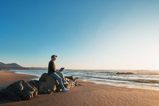 silhouette of a person working on his laptop outdoors on the shore of the beach at sunset, profile view stock photo