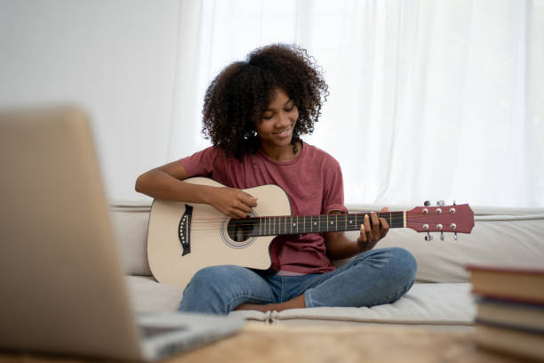 istock Focused African girl playing acoustic guitar and watching online course on laptop while practicing at home. Online training, online classes. 1472995478