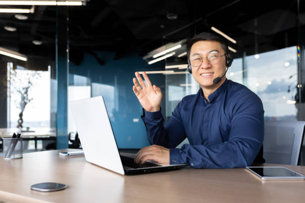 istock Man at work with headset phone and laptop successful asian man smiling and looking at camera at workplace inside office, portrait of successful businessman support worker waving hand greeting gesture 1472998109