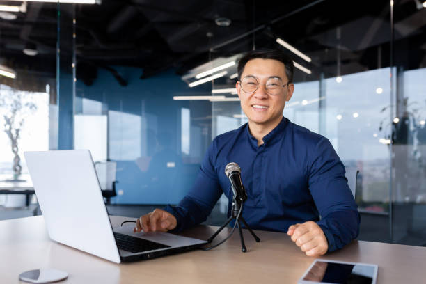 Portrait of successful asian businessman inside office, business coach teacher recording audio podcast using professional microphone, man at workplace smiling and looking at camera Portrait of successful asian businessman inside office, business coach teacher recording audio podcast using professional microphone, man at workplace smiling and looking at camera. interview seminar microphone inside of stock pictures, royalty-free photos & images