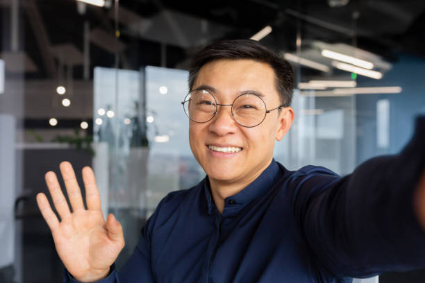 Asian man talking on video call using online communication app on phone, businessman in office taking selfie and smiling at camera, man greeting waving hand while talking with friends Asian man talking on video call using online communication app on phone, businessman in office taking selfie and smiling at camera, man greeting waving hand while talking with friends. man video call pov stock pictures, royalty-free photos & images