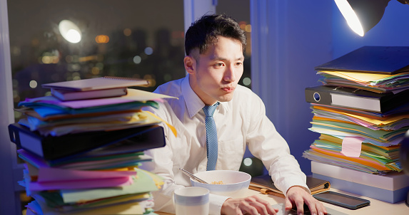 asian businessman is overtime working and hardworking at night in office - he eating instant noodle at night