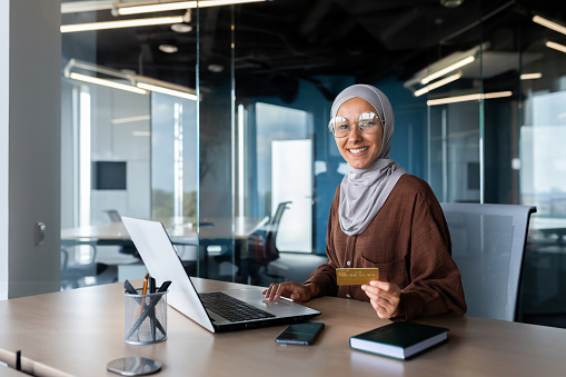 Young beautiful Muslim woman in hijab spends time online shopping. A happy woman is holding a credit card in her hands, using a laptop. He looks at the camera, smiles.
