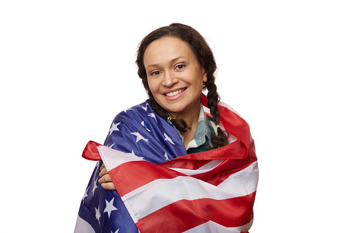 Isolated portrait on white background of charming ethnic woman, smiling a toothy smile looking at camera, wrapped in US flag, proud to be american citizen, celebrating the independence day on July 4th