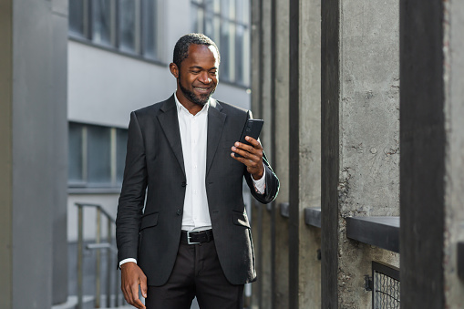 A successful African American businessman in a business suit is standing on the street near a skyscraper wearing a suit and using a mobile phone.