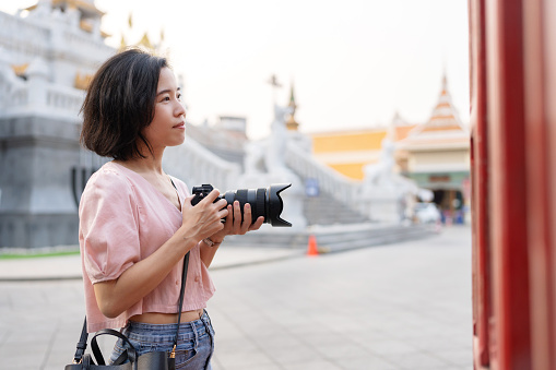 The temple is a tourist attraction that provides architecture knowledge and traditional of local people. Side view of Tourist Asian woman traveler using professional camera taking picture of temple. Holiday vacation trip during weekend activity