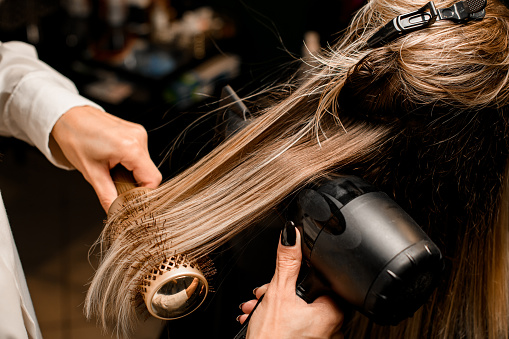Close-up of hairdo making process. Hairdresser masterfully puts long female hair with dryer and round comb