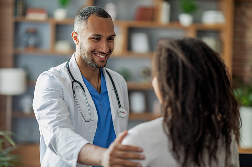 Healthcare concept. Friendly attractive middle eastern young man doctor greeting his female patient african american woman, touching her shoulder and smiling, clinic interior, closeup shot
