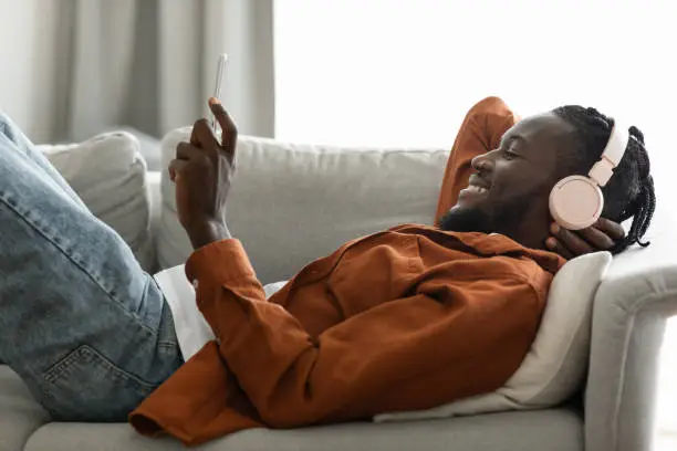 Photo of Excited african american man in wireless headphones listening to music on smartphone or watching videos, lying on sofa