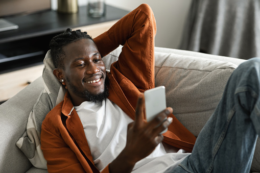 Cheerful african american man watching video on smartphone, resting and relaxing at sofa in living room interior. Video call, new app, online offer and technology at home