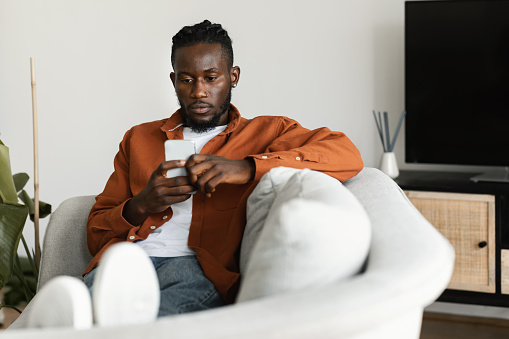 African american man sitting on couch, chatting with girlfriend or making online order via mobile app, using cellphone