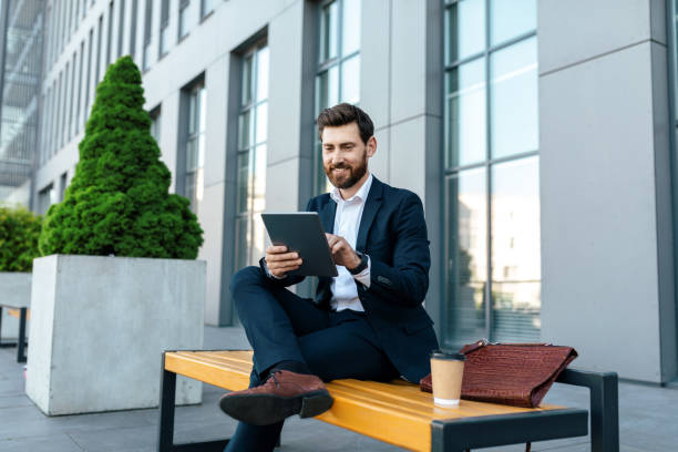 istock Smiling millennial european businessman in suit with beard typing on tablet, sit on bench, rest in city 1473016414