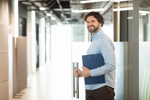 Successful Person. Portrait of confident smiling business man in shirt opening glass door standing at modern office and posing looking back at camera, holding clipboard and handle, free copy space