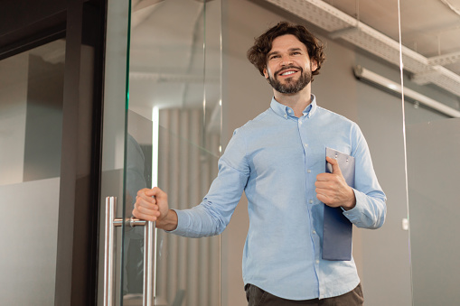 Portrait of confident smiling middle aged business man in shirt opening glass door walking in modern office and smiling, holding clipboard and handle, free copy space, low angle view