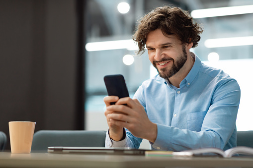People And Technology Concept. Portrait Of Smiling Businessman Using Smartphone Sitting At Desk Working In Office, Browsing New Application, Selective Focus, Blurred Background, Free Copy Space