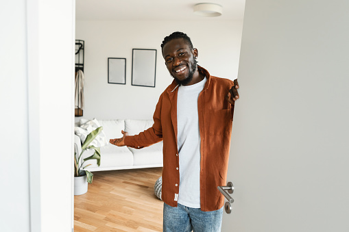 Bienvenido. Hombre afroamericano feliz invitando al visitante a entrar en su casa, de pie en la puerta del apartamento moderno photo