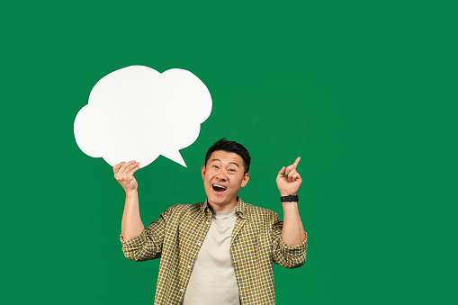 I have creative idea. Excited mature asian man holding an empty speech bubble above his head over green studio background. Mockup with space for your ad or design