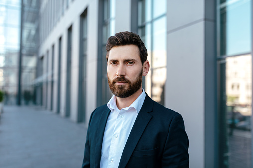 Headshot of serious attractive confident young caucasian guy with beard in suit looks at camera on street near modern office building, close up. Lifestyle of businessman, finance, startup and trade