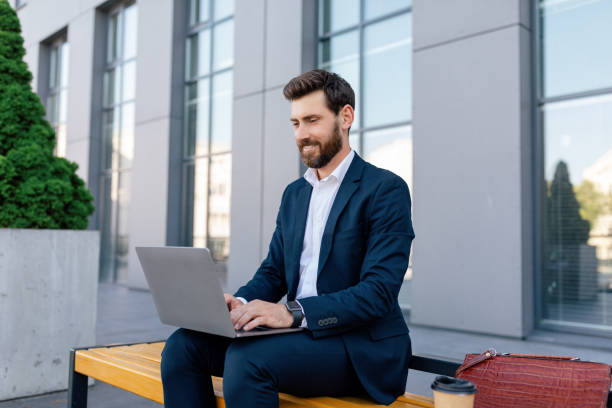 istock Cheerful attractive young caucasian guy manager with beard in suit typing on laptop, sits on bench 1473016670