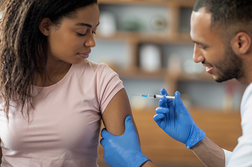 Young african american woman getting vaccinated against coronavirus, looking at male doctor handsome middle eastern man making injection in shoulder, clinic interior, closeup, copy space