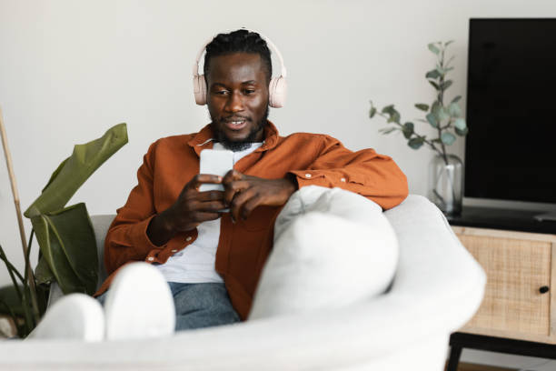 Relaxed black man watching movies on smartphone, wearing headset and using cellphone, sitting on sofa, copy space