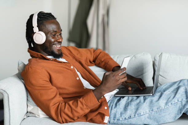 istock Smiling black man lying on couch with pc, using cellphone and wearing headset, taking break from remote work 1473017271