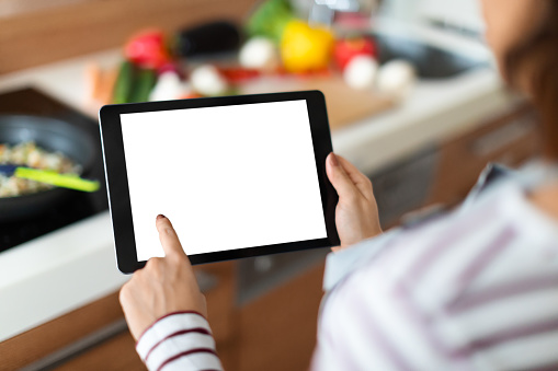 Over shoulder shot of unrecognizable woman cooking delicious healthy food at home, showing digital tablet with white blank screen, using nice cooking application, copy space, mockup