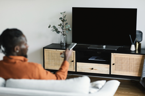 Unrecognizable black man turning on plasma flatscreen TV-set, pointing remote control at empty black TV screen, switching channels at home, mockup